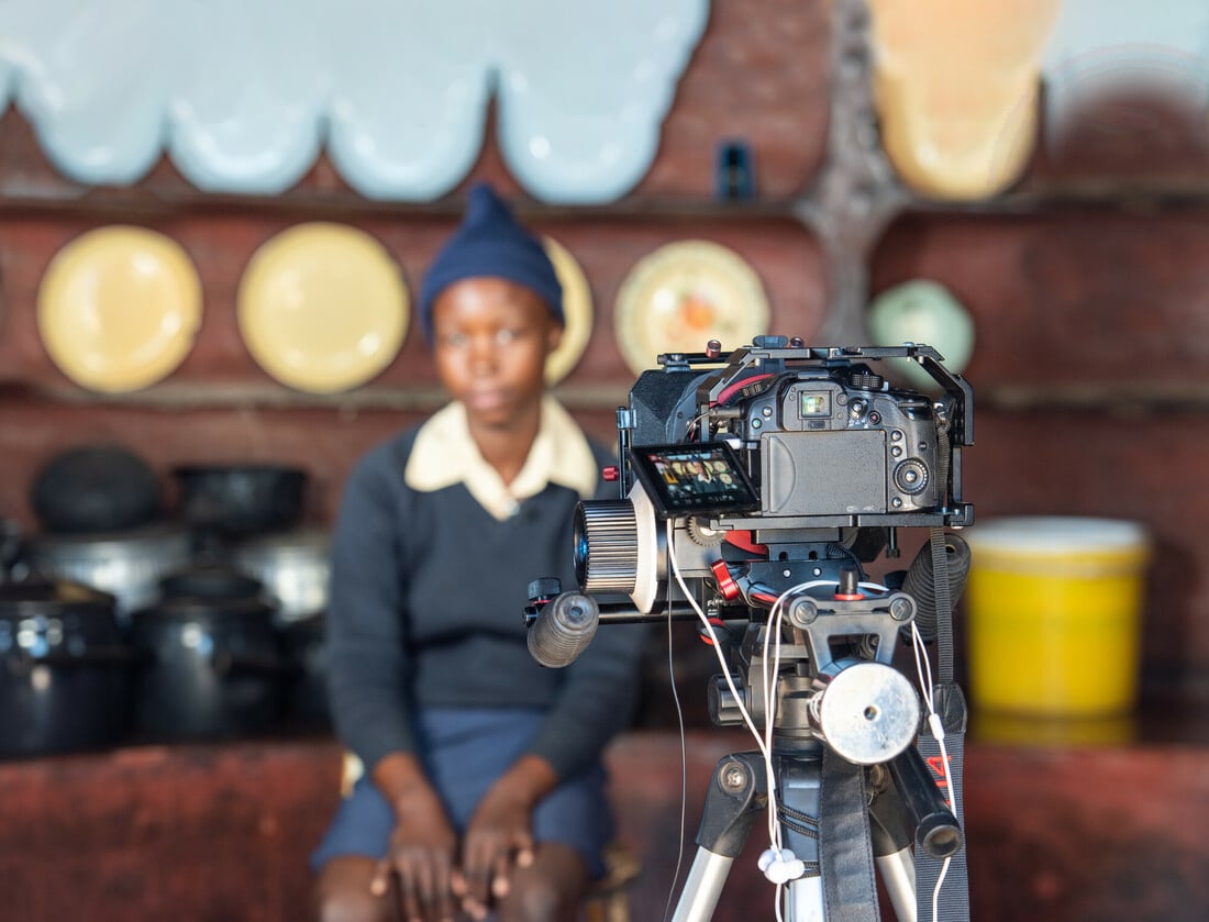 A video camera in the foreground is pointed toward Thembinkosi, a young woman in Zimbabwe telling her story.