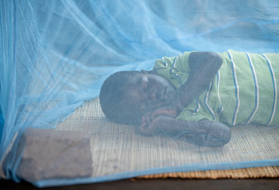 Boy in Zimbabwe sits on bed and looks up from underneath a mosquito net.