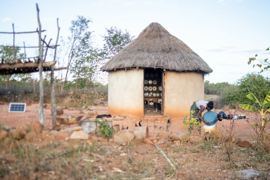 Photograph of a small hut with a young woman doing chores outside.