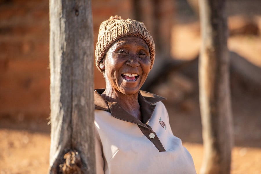 A woman grins at the camera. She stands between two tree trunks in rural Zimbabwe.