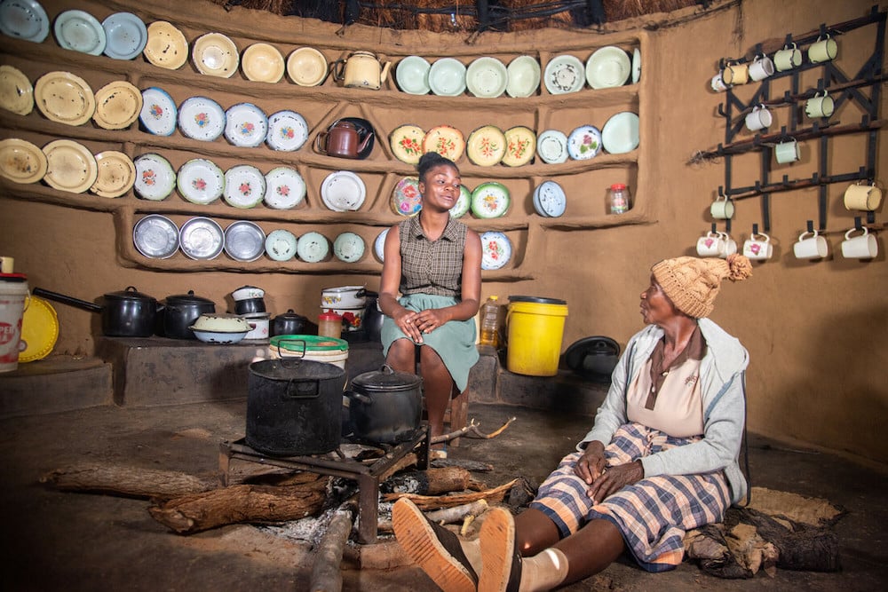 Journalist photographs young woman sitting on chair in a kitchen talking to older woman, who sits on the ground. The background shows pots, colourful plates and a mug collection.